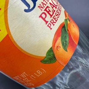 May include: Close-up of a glass jar with a label for natural peach preserves. The label features a peach illustration with green leaves and water droplets. The label also includes the text "NATURAL PEACH PRESERVES" and "NET WT. 16 OZ. (1 LB.)".