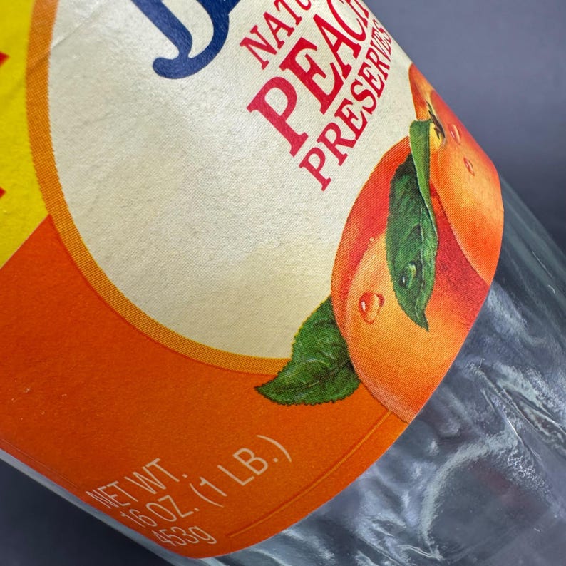 May include: Close-up of a glass jar with a label for natural peach preserves. The label features a peach illustration with green leaves and water droplets. The label also includes the text "NATURAL PEACH PRESERVES" and "NET WT. 16 OZ. (1 LB.)".