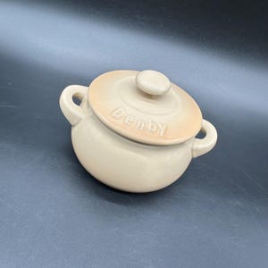 May include: A beige Denby ceramic lidded pot with two handles. The lid has a small knob and the word "Denby" is embossed on the lid. The pot is round and has a smooth finish.