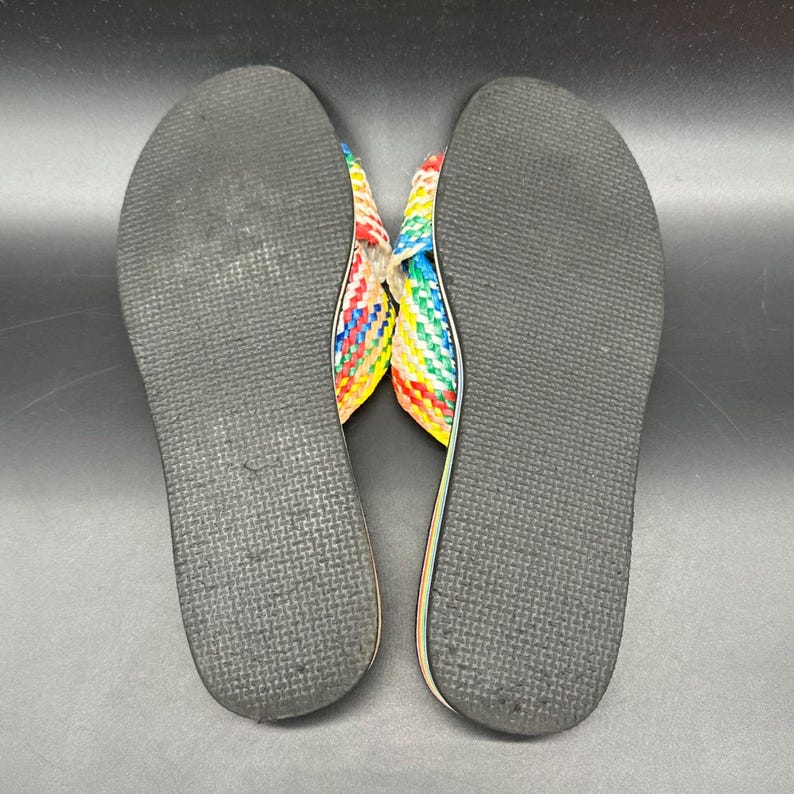 May include: A pair of colorful woven sandals with a rainbow pattern. The soles are black and textured, and the straps are woven with red, yellow, blue, and green threads. The sandals are designed for casual wear.