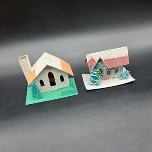 Puede incluir: Dos casas de papel en miniatura. Una tiene una chimenea beige y detalles de techo naranja, sobre una base verde. La otra tiene un techo rojo y blanco, con detalles azules y verdes, y se asienta sobre una base blanca. Ambas son decoraciones navideñas de estilo vintage.
