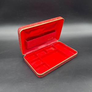 May include: Open red jewelry box with a gold trim and a patterned exterior. The interior is lined with red velvet and features compartments for storage. The box is rectangular and has a zippered closure.