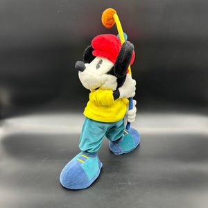 Puede incluir: Un mu&ntilde;eco de peluche de Mickey Mouse con un sombrero rojo y una camisa amarilla, sosteniendo un palo de golf con una bola en el extremo. El mu&ntilde;eco est&aacute; de pie sobre una superficie negra.