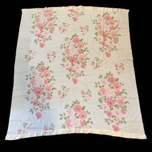May include: A white blanket with a pattern of pink roses and green leaves. The blanket has a ruffled, light yellow border. The floral design is spread across the blanket, creating a delicate and feminine aesthetic.
