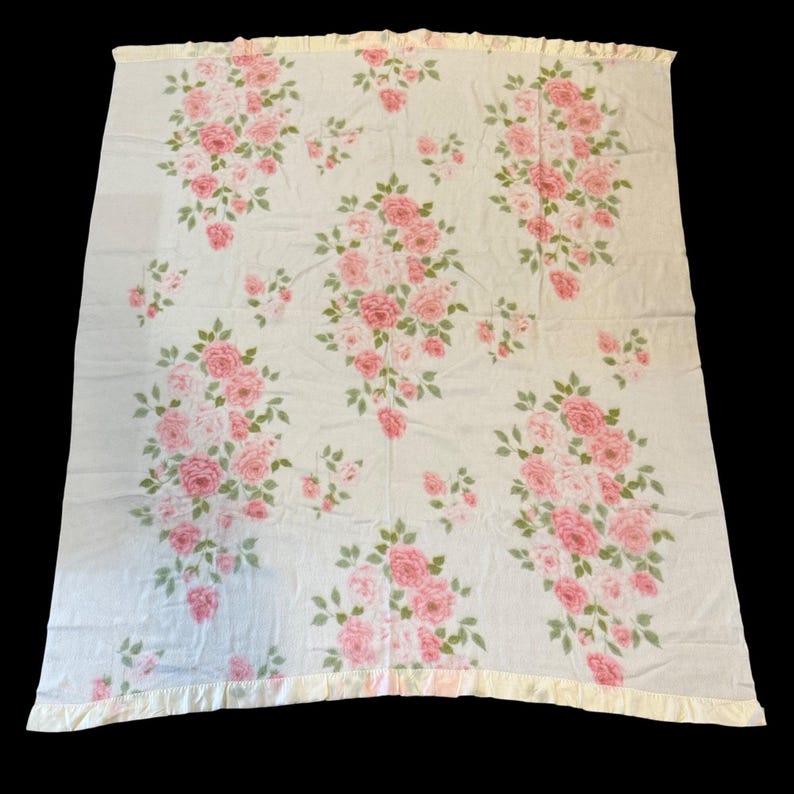 May include: A white blanket with a pattern of pink roses and green leaves. The blanket has a ruffled, light yellow border. The floral design is spread across the blanket, creating a delicate and feminine aesthetic.
