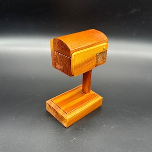 May include: A small wooden mailbox replica featuring a curved top lid and a rectangular base. The object is constructed from light brown wood with visible grain patterns and includes a small brass-colored metal hinge on the side panel.
