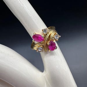 May include: A gold ring featuring three oval-cut, vibrant pink gemstones and several smaller, clear gemstones. The ring has an intricate, swirling design, with the gemstones clustered together. The ring is displayed on a white surface against a dark background.