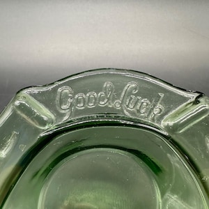 May include: A vintage green glass ashtray with the embossed text "Good Luck". The ashtray has a rounded shape with a slightly raised rim and a central depression for holding ashes. The glass has a slight transparency.