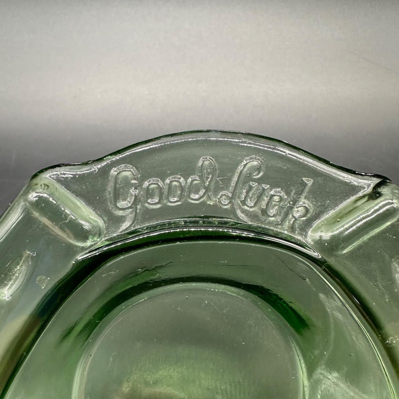 May include: A vintage green glass ashtray with the embossed text "Good Luck". The ashtray has a rounded shape with a slightly raised rim and a central depression for holding ashes. The glass has a slight transparency.