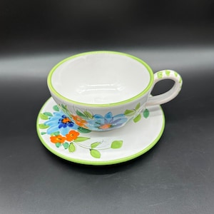 Może przedstawiać: Biały ceramiczny zestaw filiżanki i spodeczka do herbaty z zielonym rantem i kwiatowym wzorem z niebieskimi, pomarańczowymi i zielonymi kwiatami.