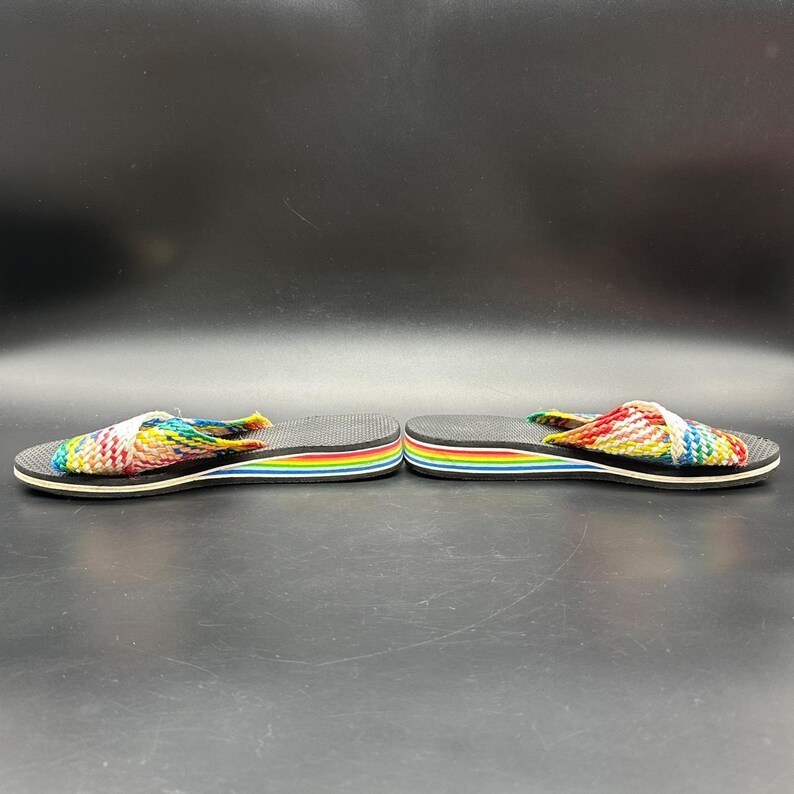 May include: A pair of colorful flip-flops with a rainbow-striped sole and a woven strap in shades of red, yellow, blue, and green. The flip-flops have a black base and a white trim.