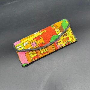 May include: A colorful, rectangular glasses case with a flap closure. The case features a vibrant pattern of buildings in shades of orange, yellow, green, and pink. The edges are trimmed with a dark gray material.