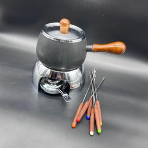 May include: A black fondue pot with a wooden handle and lid, set on a chrome base. Includes six fondue forks with wooden handles and colorful tips. The pot is designed for heating and serving melted cheese or chocolate.
