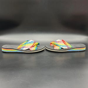 May include: A pair of rainbow-striped sandals. The sandals have a black sole with a rainbow-colored stripe around the edge. The straps are made of woven fabric in a rainbow pattern. The sandals are displayed on a dark surface.
