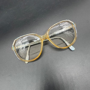 May include: Vintage eyeglasses with a light brown frame and clear lenses. The frames have a decorative pattern on the upper rims and temples. The glasses are set against a dark background.