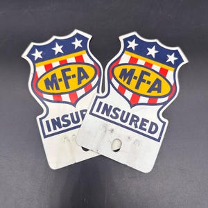 May include: Two shield-shaped metal signs featuring a blue top section with three white stars, a central yellow oval with the text M-F-A in blue, and red and white vertical stripes. The bottom white section displays the word INSURED in blue text.
