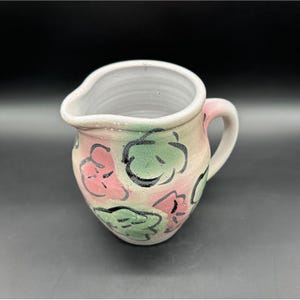 Puede incluir: Una jarra de cerámica blanca con asa, decorada con diseños florales abstractos en rosa y verde.