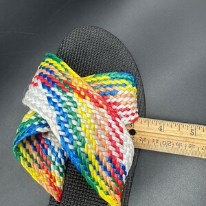 May include: Black sandal with a woven strap in a rainbow of colors. The strap is made of white, red, orange, yellow, green, and blue materials. The sandal is being measured with a ruler.