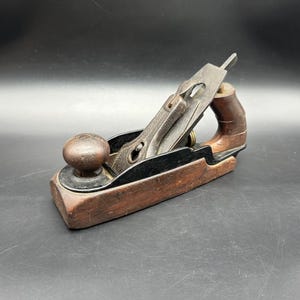 May include: A vintage wooden hand plane with a dark finish. The plane features a metal blade and lever cap, along with a wooden handle and knob. The tool is designed for woodworking and shaping materials.