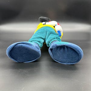 Puede incluir: Un juguete de peluche de Mickey Mouse con un atuendo azul, verde, amarillo y blanco. El juguete est&aacute; de pie sobre sus pies, con los brazos a los lados. El juguete est&aacute; mirando hacia adelante, pero la imagen se toma desde abajo, por lo que el espectador est&aacute; mirando hacia arriba a los pies del juguete.
