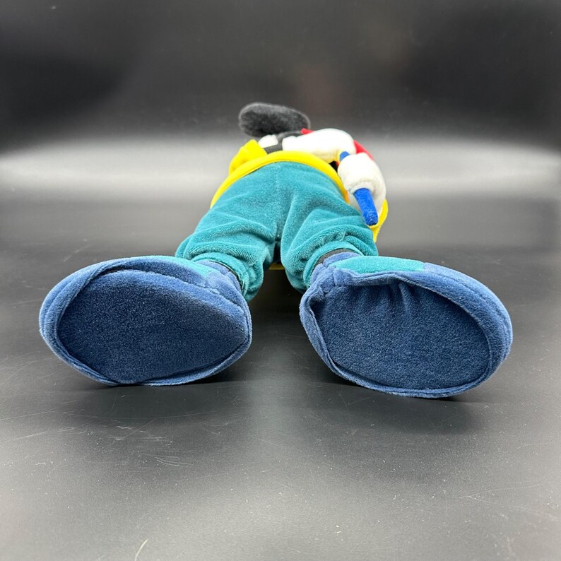 Puede incluir: Un juguete de peluche de Mickey Mouse con un atuendo azul, verde, amarillo y blanco. El juguete est&aacute; de pie sobre sus pies, con los brazos a los lados. El juguete est&aacute; mirando hacia adelante, pero la imagen se toma desde abajo, por lo que el espectador est&aacute; mirando hacia arriba a los pies del juguete.