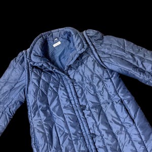 May include: A blue quilted jacket featuring a diamond-stitched pattern, a high collar, and long sleeves with gathered shoulders. The front closure consists of three fabric-covered toggle buttons. A small white rectangular label is visible at the neck.