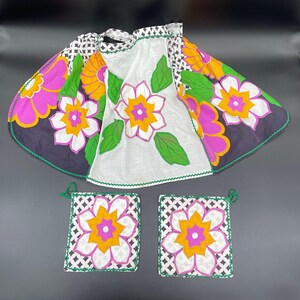 May include: A vintage floral apron and two matching pot holders. The apron features a white center with a large flower design and a black and white patterned border with large pink, yellow, and green flowers. The pot holders have the same floral design.