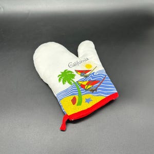May include: White oven mitt with a colorful beach scene. The mitt features a palm tree, sailboats, and the word "California." The bottom edge is trimmed in red, and the mitt has a loop for hanging.
