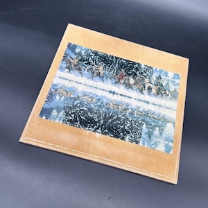 May include: A square, tan coaster with a detailed winter scene featuring riders on horseback and a snowy landscape. The artwork is centered on the coaster, surrounded by a stitched border.