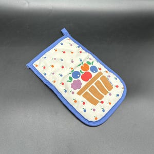 May include: A white oven mitt with a blue border and a loop for hanging. The mitt features a colorful illustration of a basket filled with fruit, including blueberries, oranges, and cherries. Small red and blue fruit designs are scattered across the white background.