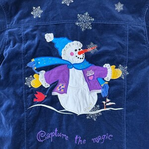 May include: Navy blue corduroy jacket with an embroidered snowman design. The snowman wears a blue hat and scarf, a purple jacket, and yellow mittens. The phrase "Capture the magic" is embroidered in purple.