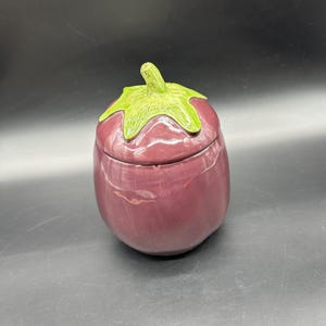 May include: A ceramic eggplant-shaped container with a lid. The container is a deep purple color, with a green stem and leaf design on the lid. The container is likely used for storage or decoration.