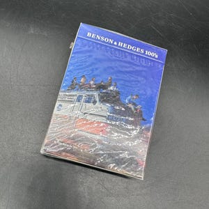 Puede incluir: Una caja de cigarrillos rectangular Benson & Hedges 100's con un gráfico azul y blanco. El gráfico representa a personas en un barco. La caja está envuelta en plástico transparente.