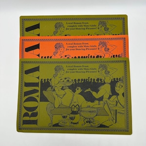 May include: Three rectangular placemats with the word "ROMA" in large black letters. The placemats are green and orange, with illustrations of Roman figures and text that reads "A real Roman Feast, complete with Slave Girls for your Dancing Pleasure!"