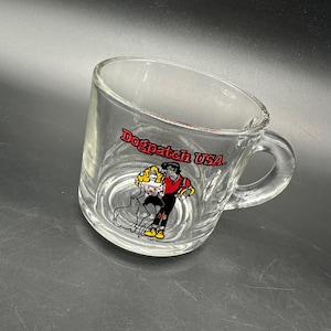 May include: Clear glass mug with a handle. The mug features a colorful graphic of a cartoon couple dancing with a dog, and the words "Dogpatch USA" in red. The mug is likely vintage and collectible.