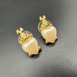 May include: A pair of gold-tone earrings featuring a rabbit holding a heart-shaped pearl. The earrings are a classic and elegant design.