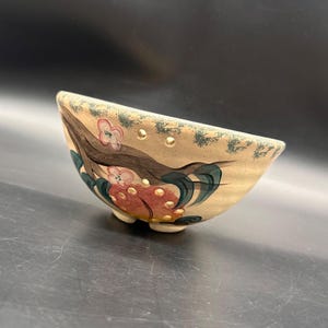 以下が含まれることがあります： ピンクの花と緑の葉が描かれた茶色の木の枝の手描きデザインが施されたベージュの陶器製ボウル。ボウルの縁の近くには小さな円形の穴が並んでおり、3つの小さな脚で支えられています。表面には斑点状のテクスチャがあります。