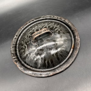 May include: A round, domed metal lid featuring a mottled black and dark gray speckled finish. The lid has a raised, arched handle centered on the top and a wide, flat rim with visible signs of wear and oxidation along the outer edge.