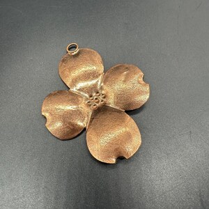 以下が含まれることがあります： 4枚の花びらの花の形をした銅色のペンダント。ペンダントはテクスチャ加工された表面と、吊り下げ用の小さなループが上部にあります。花の中心には、小さな丸いディテールが集まっています。