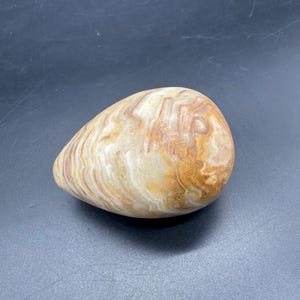 May include: A smooth, egg-shaped stone with a polished surface. The stone features a blend of beige, brown, and reddish-orange hues in a swirling pattern. The natural stone is a decorative item.