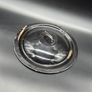 May include: Black enamel pot lid with a metal handle. The lid has a slightly raised rim and shows signs of wear.