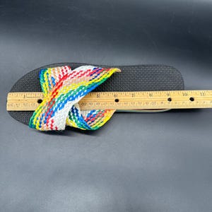 May include: Black flip-flop with a woven strap in a rainbow pattern. The flip-flop is measured with a wooden ruler. The strap is made of red, yellow, green, blue, and white woven material.