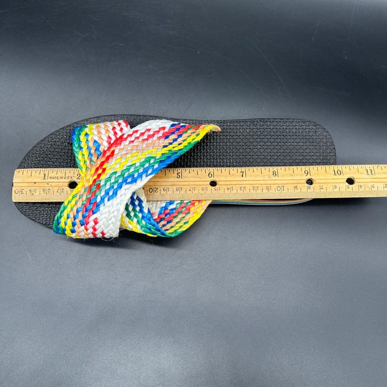 May include: Black flip-flop with a woven strap in a rainbow pattern. The flip-flop is measured with a wooden ruler. The strap is made of red, yellow, green, blue, and white woven material.