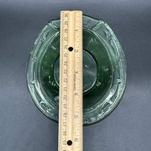 May include: A clear green glass bowl with a decorative rim, approximately 6 inches in diameter, is shown from a top-down perspective. A wooden ruler is placed next to the bowl for scale. The bowl has a circular interior and a slightly textured surface.
