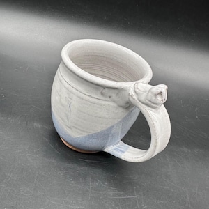 May include: A handcrafted ceramic mug with a distinctive handle. The mug has a light grey and blue colour scheme, a textured surface, and a rounded form. The handle has a decorative feature. Suitable for coffee, tea, or other drinks.