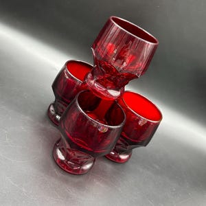 May include: A set of four ruby red vintage cordial glasses. The glasses have a faceted design on the base and a smooth rim. The glasses are stacked together, showcasing their rich color and elegant shape.