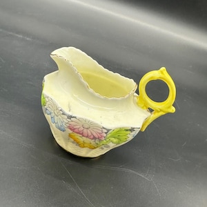 Puede incluir: Un pequeño creamer blanco con un asa amarilla y un diseño floral. El creamer tiene un borde festoneado y un borde dorado.