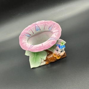 Könnte beinhalten: Ein rosafarbener und weißer Keramik-Blumentopf mit einem skurrilen Gesicht, das hinter der Blume hervorlugt. Der Topf ist in Form einer Windeblume mit einer kleinen Öffnung zum Pflanzen geformt.