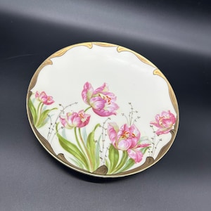 May include: A white porcelain plate with a gold rim and a floral design featuring pink tulips and green leaves.