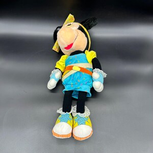 Peut inclure: Un jouet en peluche de Minnie Mouse portant une tenue bleue et jaune avec un bandeau jaune. Elle a des oreilles noires et un nez noir. Elle porte des baskets blanches avec des accents jaunes.